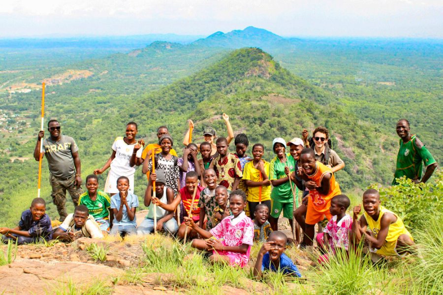 A large group stands on top of a mountain on a sunny day and poses for a photo